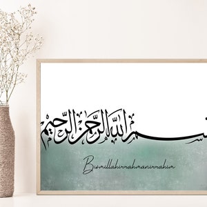 May include: A framed print featuring black Arabic calligraphy on a white background. The calligraphy reads "Bismillahirrahmanirrahim" in English. The background is a light blue watercolour wash.