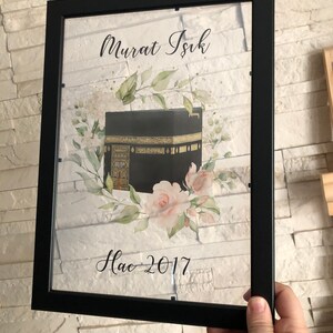 Puede incluir: Una impresi&oacute;n enmarcada con una imagen en negro y dorado de la Kaaba, rodeada de flores de acuarela. El texto "Murat Isik Hac 2017" est&aacute; impreso debajo de la imagen.
