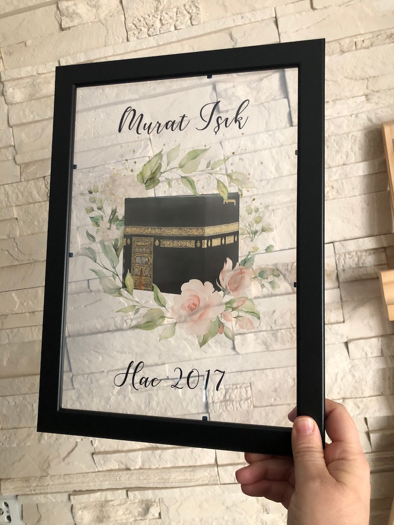 Puede incluir: Una impresi&oacute;n enmarcada con una imagen en negro y dorado de la Kaaba, rodeada de flores de acuarela. El texto "Murat Isik Hac 2017" est&aacute; impreso debajo de la imagen.