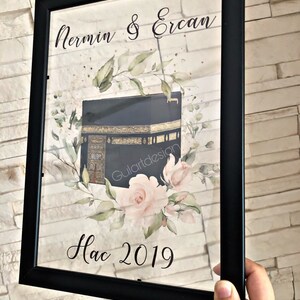 Puede incluir: Una impresi&oacute;n enmarcada con fondo negro que presenta una ilustraci&oacute;n de acuarela de la Kaaba, un edificio en La Meca, Arabia Saudita, con rosas rosadas y hojas verdes. El texto "Nermin & Ercan Hac 2019" est&aacute; impreso debajo de la ilustraci&oacute;n.