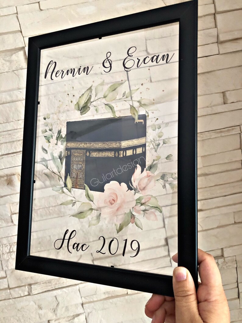 Puede incluir: Una impresi&oacute;n enmarcada con fondo negro que presenta una ilustraci&oacute;n de acuarela de la Kaaba, un edificio en La Meca, Arabia Saudita, con rosas rosadas y hojas verdes. El texto "Nermin & Ercan Hac 2019" est&aacute; impreso debajo de la ilustraci&oacute;n.