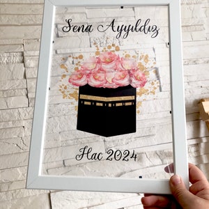 Puede incluir: Imagen enmarcada en blanco con una imagen negra y dorada de la Kaaba, rodeada de flores rosas. El texto "Sena Ayydlz Hac 2024" est&aacute; escrito en cursiva negra.