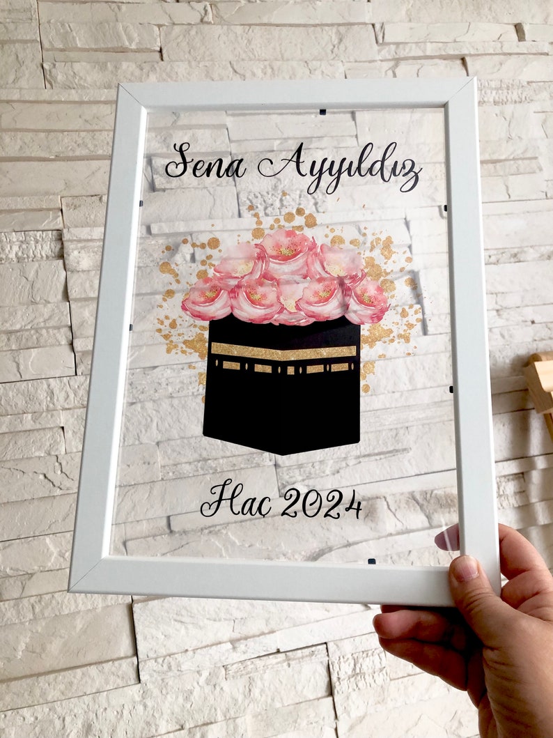 Puede incluir: Imagen enmarcada en blanco con una imagen negra y dorada de la Kaaba, rodeada de flores rosas. El texto "Sena Ayydlz Hac 2024" est&aacute; escrito en cursiva negra.