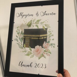 Puede incluir: Una impresi&oacute;n enmarcada con una ilustraci&oacute;n de acuarela de la Kaaba, un edificio en forma de cubo en La Meca, Arabia Saudita. La impresi&oacute;n presenta un dise&ntilde;o floral con rosas rosadas y hojas verdes. El texto "Shpejtim & Saveta Umrah 2023" est&aacute; impreso debajo de la ilustraci&oacute;n.