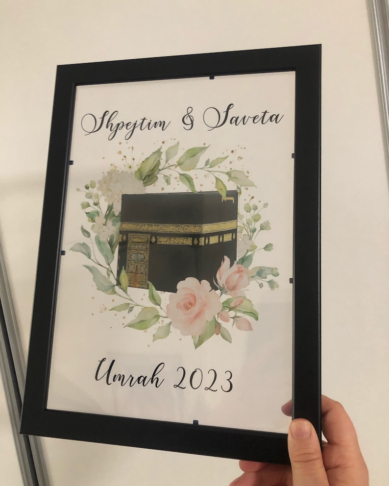 Puede incluir: Una impresi&oacute;n enmarcada con una ilustraci&oacute;n de acuarela de la Kaaba, un edificio en forma de cubo en La Meca, Arabia Saudita. La impresi&oacute;n presenta un dise&ntilde;o floral con rosas rosadas y hojas verdes. El texto "Shpejtim & Saveta Umrah 2023" est&aacute; impreso debajo de la ilustraci&oacute;n.