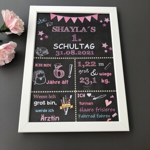 Puede incluir: Una pizarra negra con marco blanco, decorada con detalles en rosa y azul. La pizarra dice "Shaylas 1. Schultag 31.08.2021" e incluye detalles sobre la edad, la altura y el peso del ni&ntilde;o. Tambi&eacute;n enumera las actividades que le gustan al ni&ntilde;o, como hacer gimnasia, peinarse y andar en bicicleta.