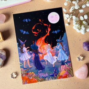 Peut inclure: Une impression d'art rectangulaire représentant quatre personnages en robe et chapeau pointu dansant autour d'un feu de joie orange vif sous un ciel nocturne avec une pleine lune et des chauves-souris. Le texte Sandra Tews est visible.