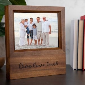 May include: A wooden picture frame with a family photo. The frame is a warm brown color and has the words "live love laugh" engraved on the front. The photo shows a family of six on a beach. The frame is sitting on a dark surface.