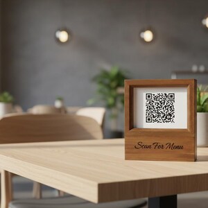May include: A wooden table sign displaying a QR code and the text "Scan For Menu". The frame is made of light brown wood. The background shows a restaurant setting with tables and chairs.