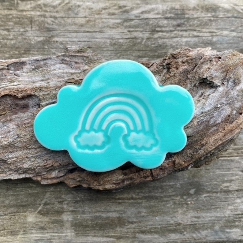 Op de afbeelding: Een turquoise blauwe keramische wolk met een wit regenboog- en wolkenontwerp.