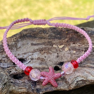 Puede incluir: Pulsera de cord&oacute;n trenzado rosa con cuentas rojas y transparentes y un dije de estrella de mar rosa.