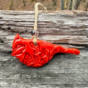 May include: A vibrant red ceramic bird ornament with a detailed floral pattern. The bird is suspended by a natural twine cord and a decorative bead. The ornament is designed for hanging and adds a touch of nature-inspired decor.