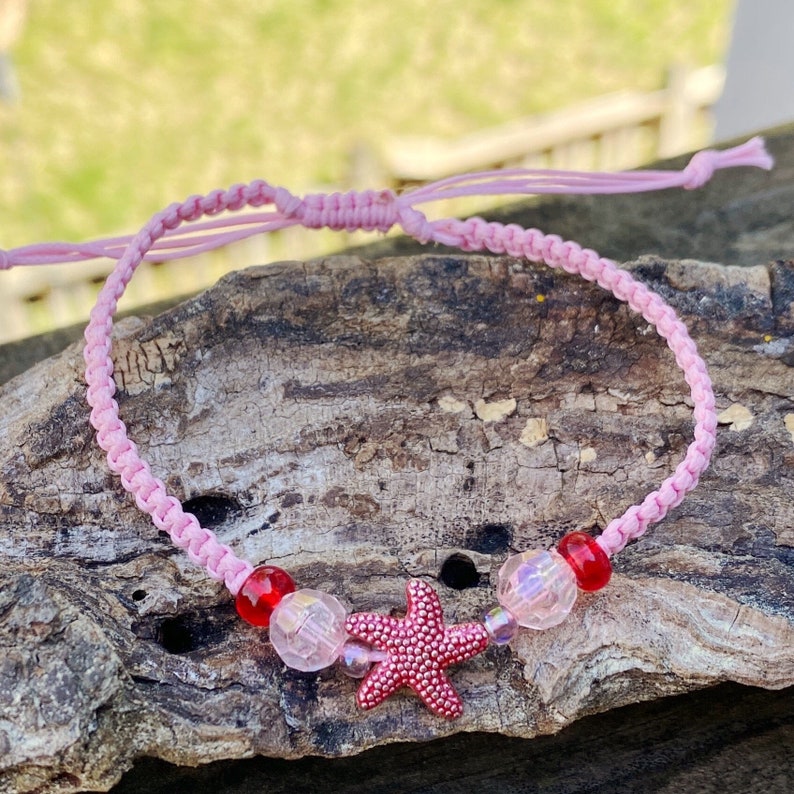 Puede incluir: Pulsera de la amistad trenzada rosa con cuentas rojas y transparentes y un dije de estrella de mar rosa.