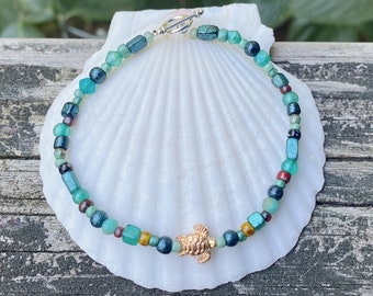 Puede incluir: Un brazalete de cuentas verdes y doradas con un dije de tortuga dorada. El brazalete está sobre una concha blanca.