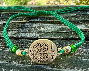 May include: A green macrame bracelet with a silver-colored circular charm featuring a tree design. The bracelet has green and gold beads. The background is a weathered wooden surface.