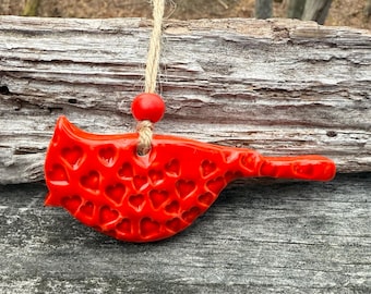 Puede incluir: Adorno de cerámica roja brillante con forma de pájaro, decorado con recortes en forma de corazón. Cuelga de un cordón de hilo natural y una cuenta roja. El adorno se presenta sobre un fondo de madera envejecida, lo que sugiere una estética rústica o artesanal.