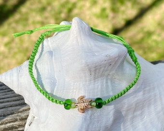 Puede incluir: Un brazalete de cuerda verde con un dije de tortuga dorada y cuentas de vidrio verde.