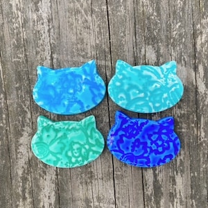 Puede incluir: Cuatro posavasos con forma de gato en tonos azules y turquesas. Cada posavasos tiene un diseño único veteado o estampado. Los posavasos están dispuestos sobre una superficie de madera envejecida.