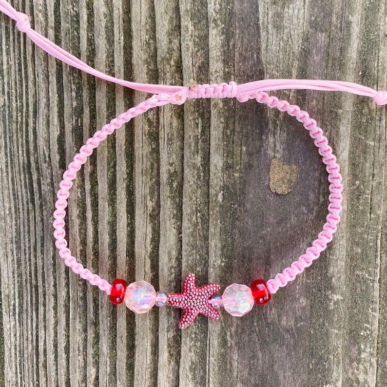 Puede incluir: Pulsera trenzada rosa con cuentas rojas y transparentes y un dije de estrella de mar rosa.