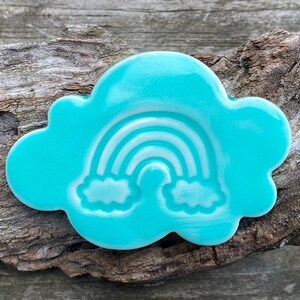 Op de afbeelding: Een turquoise blauwe keramische wolk met een witte regenboog en wolken die erop zijn gegraveerd.