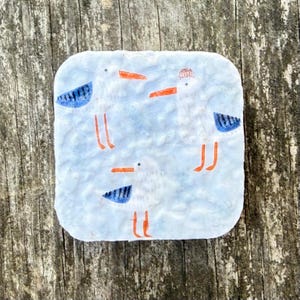 Op de afbeelding: Vierkante keramische onderzetter met een lichtblauwe achtergrond en drie cartoonmeeuwen. De meeuwen hebben witte lichamen, oranje snavels en poten en blauwe vleugelaccenten. Eén meeuw draagt een rood-witte hoed.