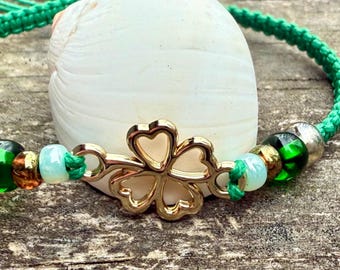 May include: A green cord bracelet with gold four-leaf clover charm and green, brown, and white beads.