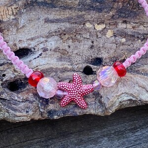 Puede incluir: Collar de cord&oacute;n trenzado rosa con cuentas rojas y transparentes y un dije de estrella de mar rosa.