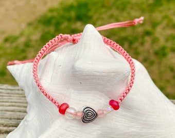 Puede incluir: Pulsera de macramé rosa con un dije de corazón plateado y cuentas rojas y transparentes. La pulsera ajustable se muestra sobre una superficie blanca con un fondo verde. Una joya delicada y encantadora.