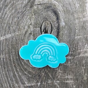 Op de afbeelding: Een turquoise blauwe wolkvormige koekjesuitsteker met een wit regenboog- en wolkenontwerp.