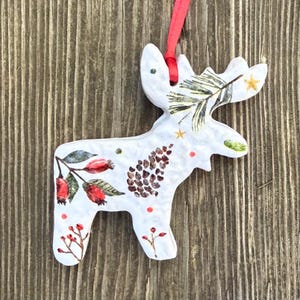 May include: White ceramic moose ornament with red ribbon for hanging. The moose is decorated with a pine cone, sprigs of berries, and a pine branch. The ornament is approximately 4 inches tall.