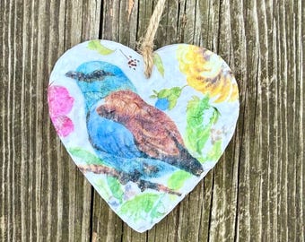 Op de afbeelding: Hartvormige ornament met een blauwe en bruine vogel op een tak, omringd door roze en gele bloemen en groene bladeren. Het ornament hangt aan een natuurlijke touwtje, tegen een achtergrond van verweerd hout.
