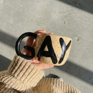 Può includere: Una tazza di ceramica maculata con la parola "GAY" in grassetto nero. La tazza ha un manico nero e uno sfondo beige con piccole macchie marroni e arancioni. La tazza è tenuta da una persona che indossa un maglione beige.
