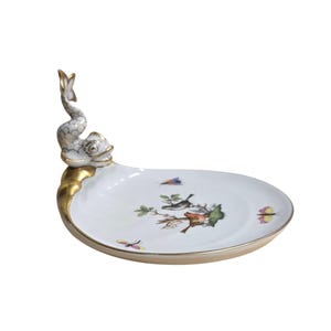 May include: White porcelain serving dish with a gold rim and a dolphin-shaped handle. The dish features a painted design of birds, butterflies, and floral elements. A decorative piece for serving or display.
