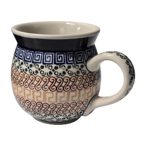 May include: A round ceramic mug featuring a dark blue rim and a handle decorated with small dark blue floral patterns. The body displays horizontal bands of geometric designs, including Greek key patterns in blue and tan, and rows of dark blue dots.