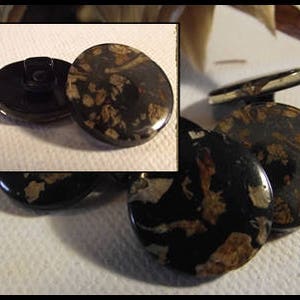 May include: Several black buttons with a speckled pattern in brown and gold tones. The buttons are circular and have a shiny surface. One button features a shank on the reverse, while others are flat.