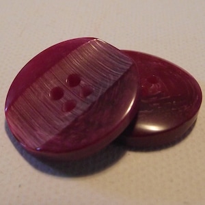 May include: Two maroon oval buttons. Each button has four holes and a diagonal textured design on one side. The buttons are glossy and appear to be made of a hard plastic or resin material. The buttons are likely for sewing or crafts.