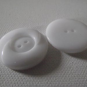 Könnte beinhalten: Zwei weiße, glänzende Knöpfe. Ein Knopf hat ein spiralförmiges Design um die beiden Knopflöcher, der andere ist eine einfache ovale Form mit zwei Löchern. Die Knöpfe liegen auf einer strukturierten, cremefarbenen Oberfläche.