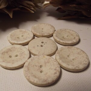May include: Seven off-white, round buttons with two or three holes each. The buttons have a slightly textured surface with subtle brown marbling. The buttons are arranged in a circular pattern. Each button is approximately 2.5 cm in diameter.