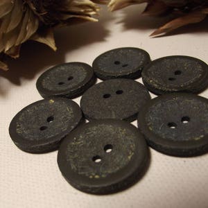 May include: Seven dark grey buttons with two holes each. The buttons have a textured surface and a slightly oval shape. The buttons are arranged in a circular pattern on a light pink surface.