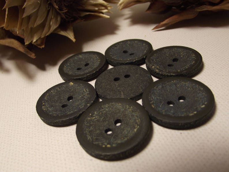 May include: Seven dark grey buttons with two holes each. The buttons have a textured surface and a slightly oval shape. The buttons are arranged in a circular pattern on a light pink surface.