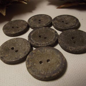 May include: Seven round, grey buttons with two holes each. The buttons have a textured surface and are arranged in a slightly overlapping pattern. The buttons are made of a natural material.