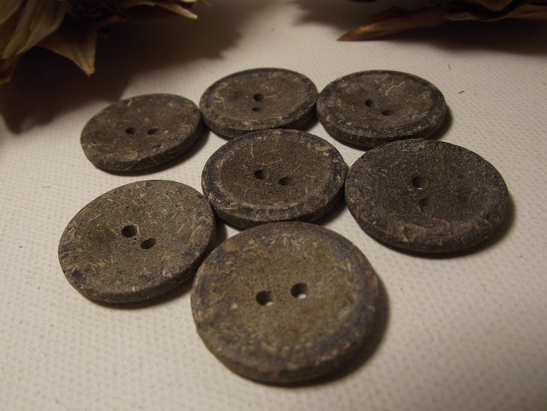 May include: Seven round, grey buttons with two holes each. The buttons have a textured surface and are arranged in a slightly overlapping pattern. The buttons are made of a natural material.