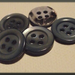 Puede incluir: Colección de cinco botones gris oscuro, cada uno con cuatro agujeros. Un botón tiene un acabado metálico plateado. Los botones están dispuestos sobre una superficie de color beige claro, mostrando sus diseños y variaciones de color.