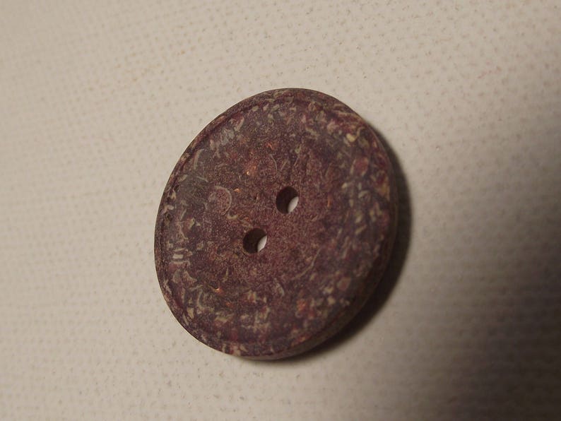 May include: A close-up of a round, dark brown button with two holes. The button has a textured surface with lighter brown and tan speckles. Suitable for sewing projects and crafts.