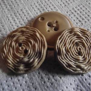 Puede incluir: Tres botones beige de diferentes diseños. Dos botones tienen un patrón en espiral con dos agujeros, mientras que el tercero es un botón redondo más grande con cuatro agujeros. Los botones están sobre una superficie blanca texturizada.