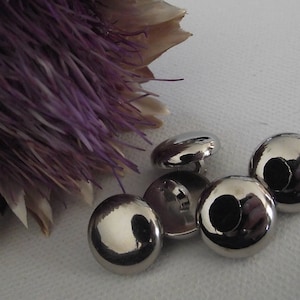 Puede incluir: Un conjunto de botones brillantes de color plateado, con forma abovedada. Los botones están agrupados sobre una superficie blanca, con una flor morada seca en el fondo. Los botones tienen una superficie reflectante.