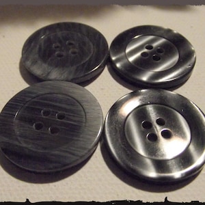 Peut inclure: Quatre gros boutons ronds avec quatre trous chacun. Les boutons sont de couleur gris foncé avec un effet marbré. Deux boutons ont un aspect métal brossé. Les boutons sont sur une surface blanche.