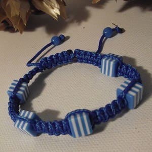 May include: A blue macrame bracelet with square, blue and white striped beads. The bracelet has an adjustable sliding knot closure and is displayed on a white surface. The bracelet is a handmade jewelry item.