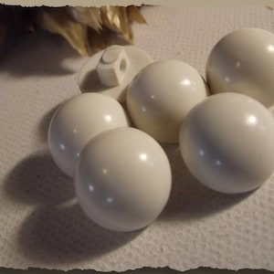 May include: Six white pearl-like buttons, some with a slight sheen, arranged on a white surface.