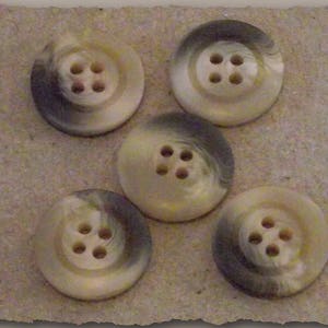 Puede incluir: Cinco botones redondos con cuatro agujeros cada uno. Los botones tienen un patrón de remolino crema y gris. Están dispuestos sobre una superficie texturizada de color marrón claro.